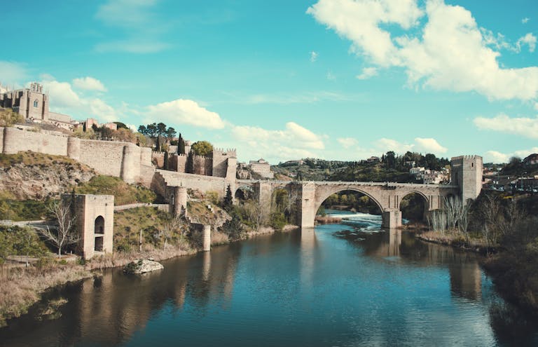 Scenic view of the Roman bridge in Toledo, Spain, reflecting over a calm river under a bright sky.