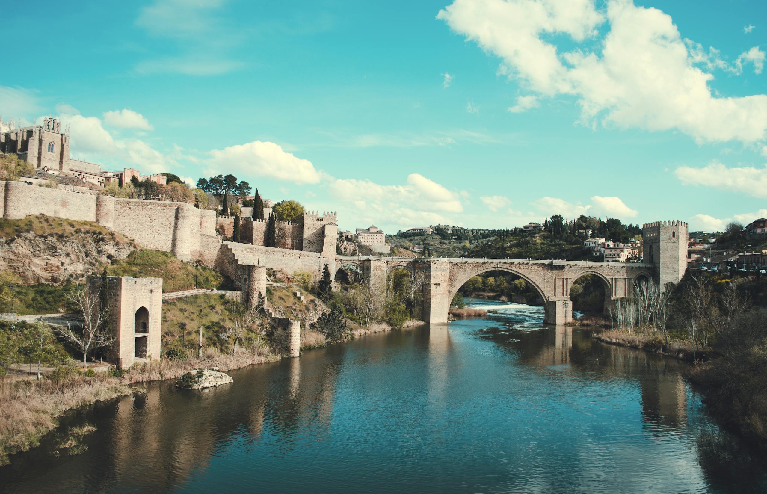 Scenic view of the Roman bridge in Toledo, Spain, reflecting over a calm river under a bright sky.